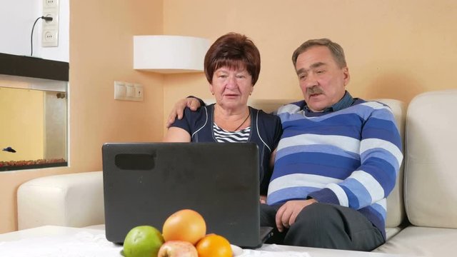 Happy senior man and a woman watching the movie on the laptop. They hug and discuss what is happening on the screen. Beautiful interior of the house