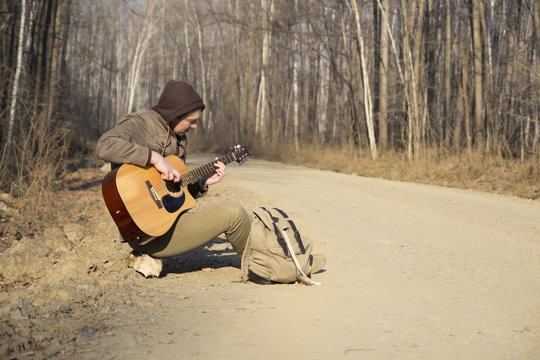 A man in the hood with a backpack sitting by the roadside and plays guitar