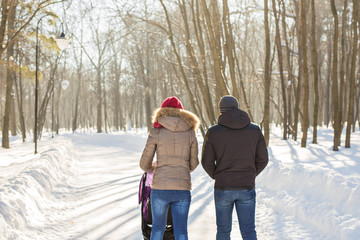 Happy young family walking in the park in winter. The parents carry the baby in a stroller through the snow.