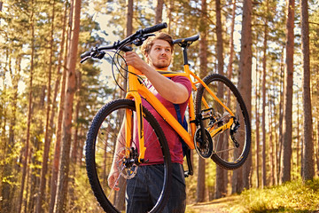 Positive sporty male holds mountain bicycle on a shoulder.