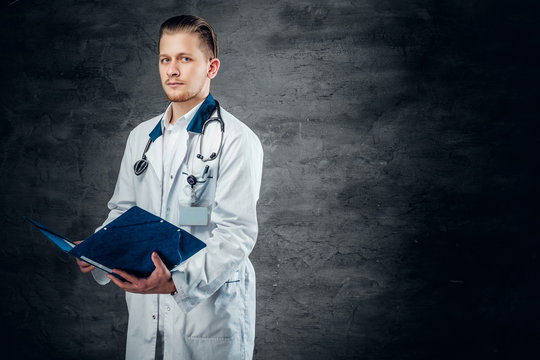 Studio portrait of young medical doctor holds blue clipboard on grey background.