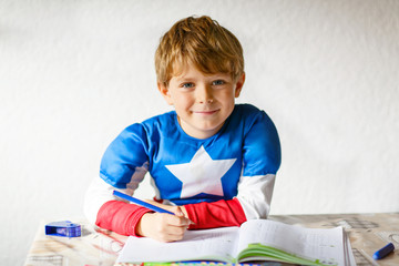 Happy school kid boy with superhero costume at home making homework