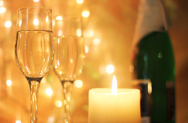 Light yellow colors Christmas greetings card or New Years Eve celebration background with an elegant arrangement with flutes and bottle of champagne and burning candles. Bokeh light garland backdrops.