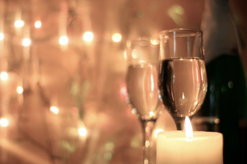 Light colors style Christmas greetings or New Years Eve celebration background with an elegant arrangement with flutes and bottle of champagne and burning candles. Bokeh light garland backdrops. 