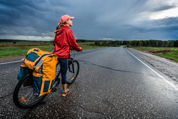 Lady hiker standing with loaded bicycle on the paved asphalt road with stormy clouds on the...