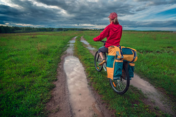 Obraz premium Young lady hiker with loaded bicycle standing on a wet rural road in the meadow