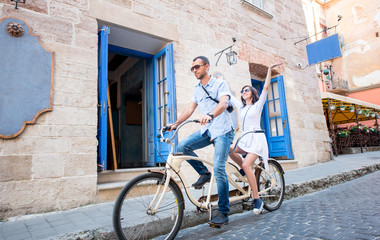 Fototapeta premium Man and woman in sunglasses riding together tandem bike on town street. The man runs a bicycle, a girl raised her hands up