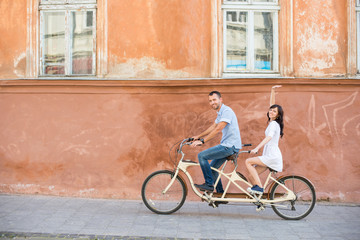 Happy couple riding on retro tandem bicycle at the street city against the background of the old orange wall with windows. The man runs a bicycle, a girl in white dress raised her hand up. Lviv