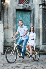 Fototapeta premium Happiness couple riding on retro tandem bicycle against the backdrop of historic building. Man in jeans and a shirt and a woman in a white dress