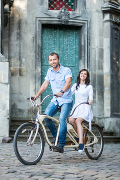 Happiness couple riding on retro tandem bicycle against the backdrop of historic building. Man in jeans and a shirt and a woman in a white dress