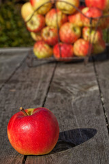 Einzelner Apfel und weitere Äpfel in einem Korb aus Draht auf einem Holztisch im Freien