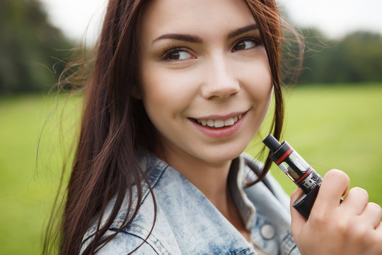 Young girl vaping.Portrait of beautifiul brunette female model smoking vape cigarette in green park outdoor.Smoke chick holding ecig vaper device in hand.Portrait of smoker with happy toothy smile