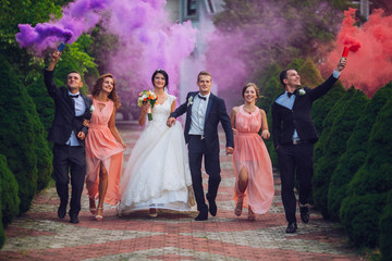 Newlyweds and their friends raise their hands up posing on the lawn. Running with smoke bombs.