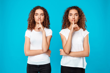 Two pretty girls twins showing keep silence over blue background.