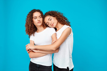 Two young pretty girls twins embracing, smiling over blue background.