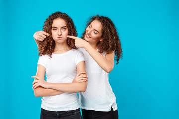 Girl smiling, touching cheeks her sister twin over blue background.
