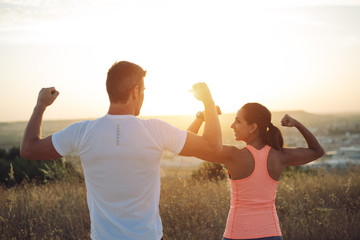 Couple of athletes flexing biceps towards the sunset. Couple of athletes celebrating running outdoor workout success. - 125597337