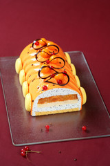 Contemporary Yule Log (layered) Cake covered with orange glaze and decorated with french macarons and chocolate loops, on burgundy background. 
