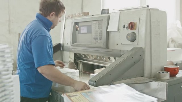 Worker in blue jacket works on cutter guillotine machine in a printing factory