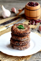 Fried bean burgers with garlic and spices on a plate. Small decorative barrel with raw red beans, garlic, parsley, fork, knife on an old wooden background. Healthy diet burgers. Rustic style. Closeup