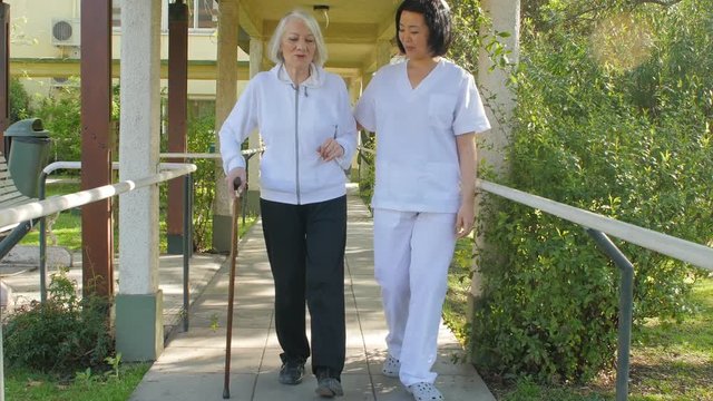 Asian nurse helping elder woman with stick in hospital garden