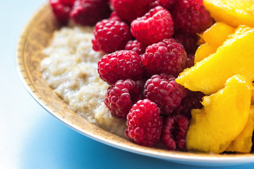 Organic oatmeal porridge in white ceramic bowl with raspberries, peaches and blueberries. Healthy breakfast - health and diet concept on the wooden table, close up.
