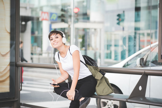 young handsome caucasian brown straight hair woman sitting on a bench at bus stop, listening music with headphones, overlooking right, smartphone handheld - technology, social network, music concept