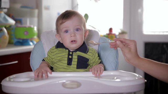 Cute smiling baby eating, sitting at baby seat at home. Mother Feeding her adorable one year old with a spoon. HD.