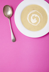A bowl of hot chicken soup with swirl of cream on a bright pink background with spoon and blank space below