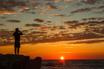 Hipster guy taking pictures of amazing landscape on mobile smart phone digital camera while standing on a rock near the sea, beautiful sunset light create background with copy space for text message