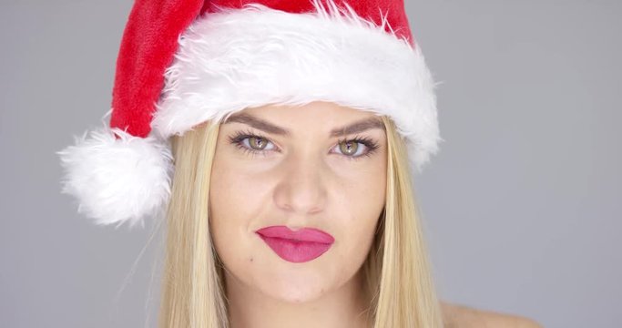 Close up portrait of beautiful blond girl in Santa Claus Hat. She posing isolated on gray backround and smiling to the camera. Her lips are painted with red lipstick.