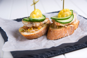 Bruschetta crostini with chopped vegetables, fried chicken, cucumber and squash on a slate plate, parchment, on a white table. Italian cuisine