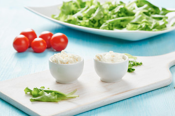 Cottage cheese in two white bowls on blue wooden table, tomatoes and salad in background