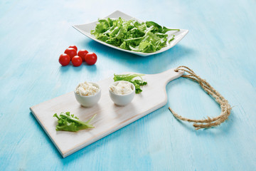 Cottage cheese in two white bowls on blue wooden table, tomatoes and salad in background
