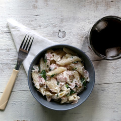 Pasta penne is cooked al dente, then mixed with sliced ham, feta cheese and basil. Dressing is made with olive oil, bit of vinegar, salt and pepper. Served with glass of soda on white rustic table.