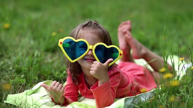 Funny little girl in big sunglasses in the shape of hearts lies on green grass and makes grimases. Beautiful little girl in glasses lies on the green grass and looks at the camera
