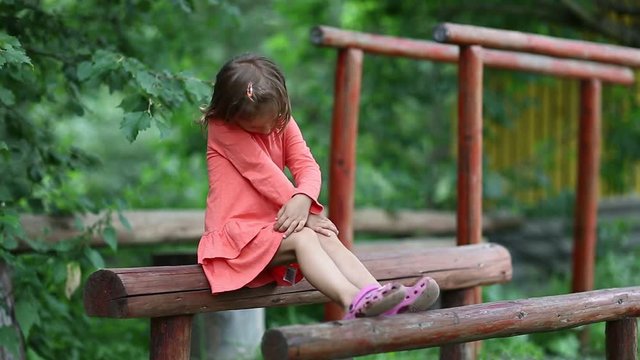 Funny little girl sits on a wooden log at a sports ground. Little girl in red dress sits on a balance beam near parallel bars and looks at the camera
