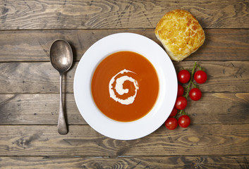 A bowl of hot soup surrounded by ripe vine tomatoes and bread roll on a rustic wooden table background