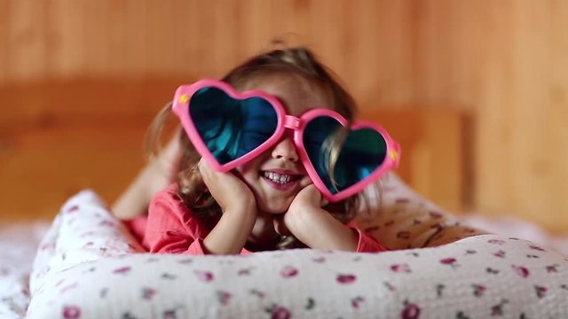 Funny girl in big glasses in the shape of hearts lies on the bed. Little girl lies on the pillow looks at the camera and shows his tongue
