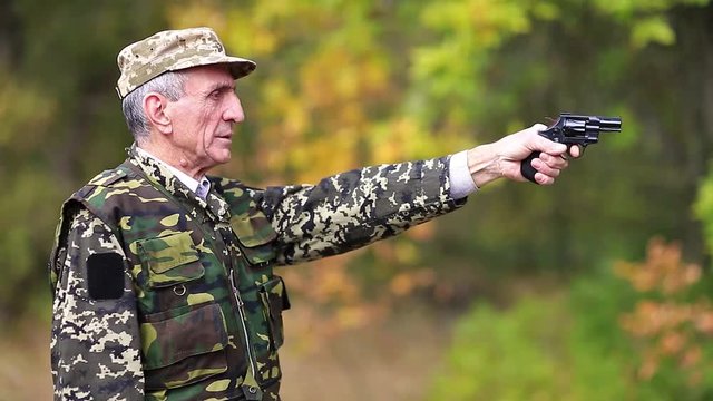 Elderly person in military uniform shoots a revolver. Retired officer at shooting range. Senior man in military uniform shoots a pistol in forest. Man with black gun. Video with sound
