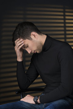 Stylish young man in a black sweater sits near blinds and thinki