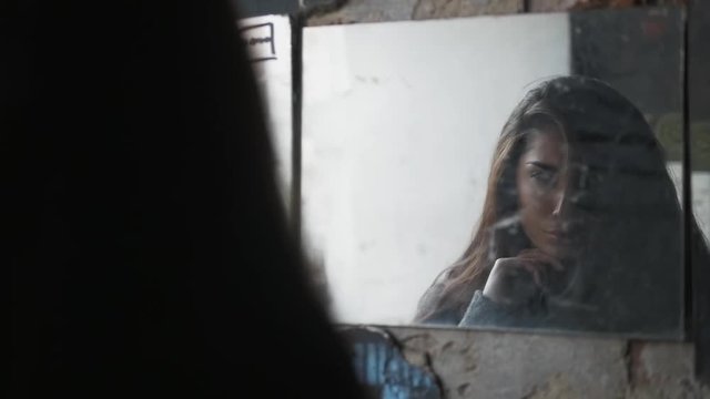 Beautiful young brunette girl with long hair looking at herself in a broken mirrors on street