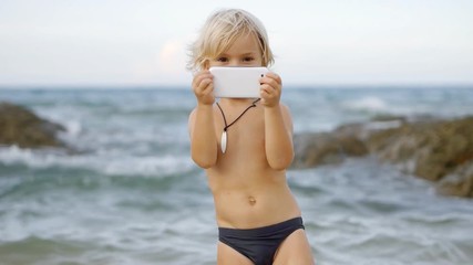 A cute little child video chats on smart phone on the beach on blue sea background. Using  a portable handheld device on summer vacation. Talking on a mobile - Powered by Adobe