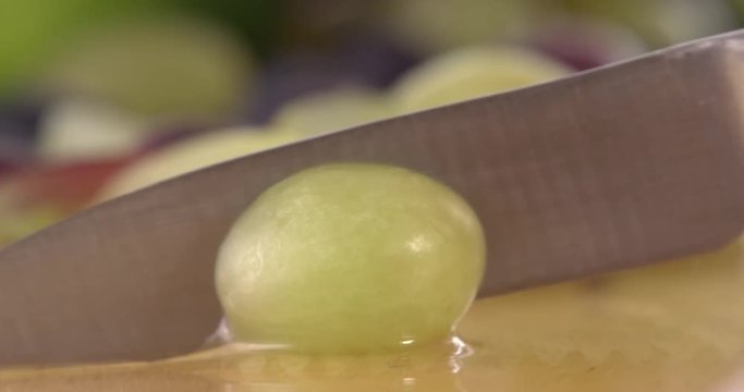 Slicing a grape in half using a sharp kitchen knife. Filmed with selection of fruit in background and on a wooden chopping board. Filmed in slow motion.