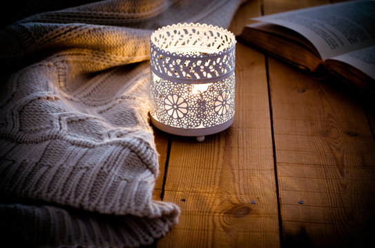 Lit candle in a lace candle holder, knitted sweater and open book on wood background