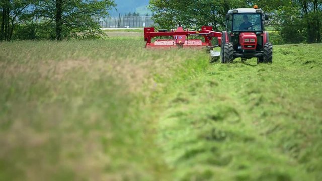 A farmer is driving his tractor across a field of grass and he is cutting grass with a grass cutting machine. Wide-angle shot.
