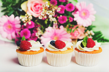 3 cupcake with butter cream and fresh berries, bouquet of pink flowers on background