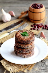 Fried bean cutlets with garlic and spices on a plate. Small decorative barrel with uncooked red beans, garlic, parsley, fork, knife on wooden background. Healthy diet cutlets. Rustic style. Closeup