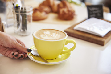 Woman’s hand and cappuccino coffee in yellow cup and saucer