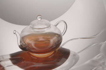 Transparent glass teapot with tea on the reflective surface on a light gray background